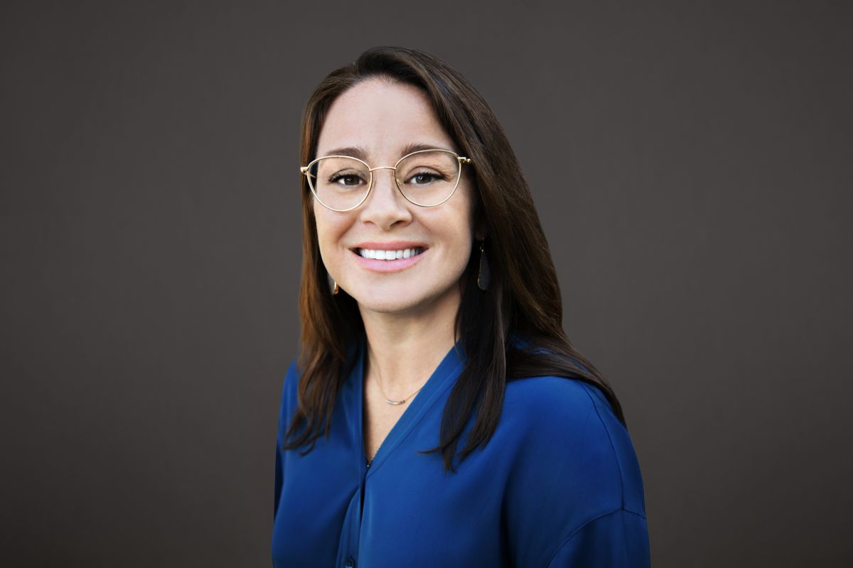 Professional headshot of Lauren Dunning, a woman with straight brown hair wearing round gold glasses, drop earrings, and a cobalt blue blouse. She is smiling and posed against a dark gray background.