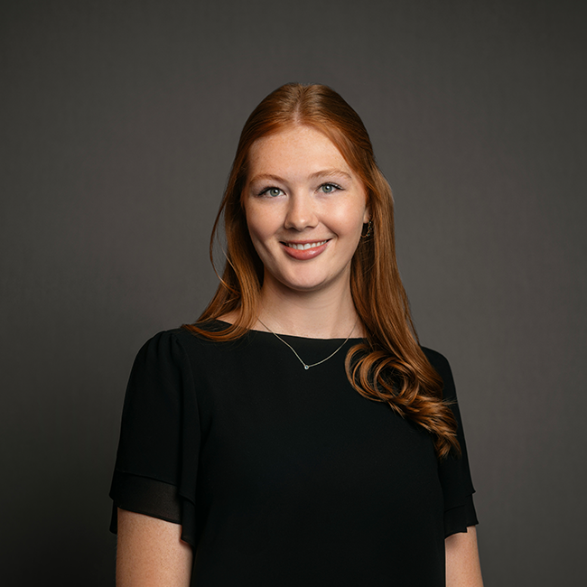 Professional headshot of Madelyn McLaughlin, a woman with long red hair styled in loose waves, wearing a black blouse and a delicate necklace. She is smiling and posed against a dark gray background.
