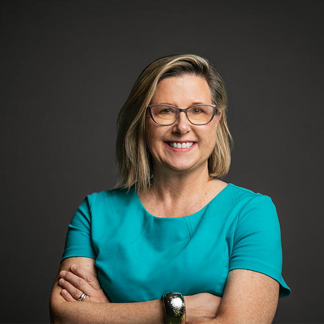 Professional headshot of Sarah Wells Kocsis, a woman with shoulder-length blonde hair and glasses, wearing a turquoise dress. She is smiling with arms crossed and posed against a dark gray background.