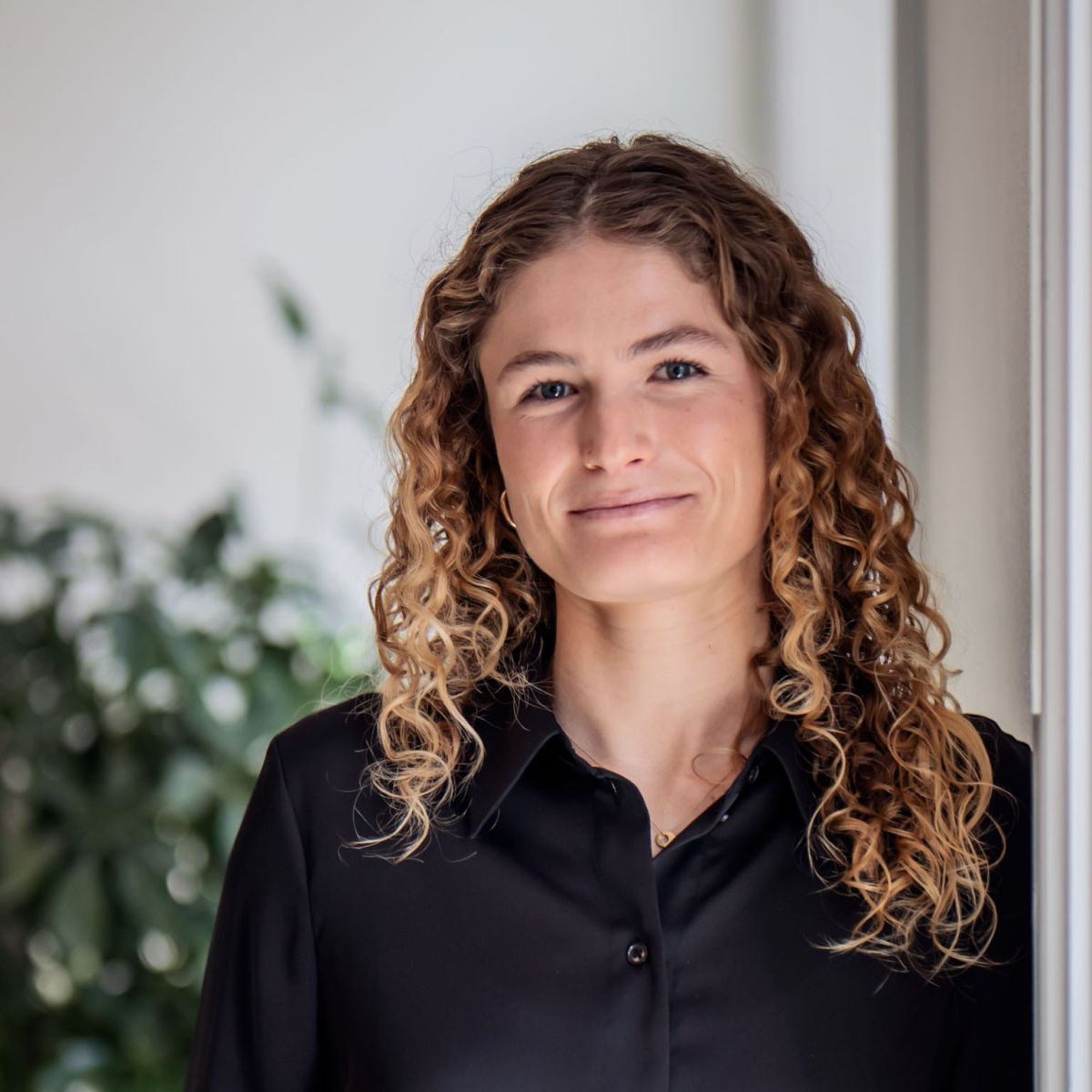 A woman with curly blonde hair wearing a black button-down shirt smiles softly while standing indoors, with greenery blurred in the background.