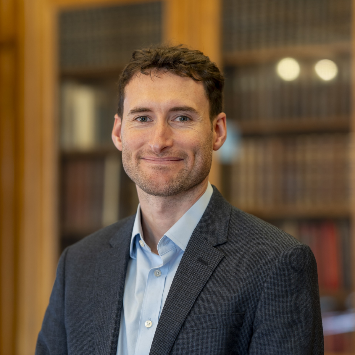 Man with short brown hair wearing a light blue shirt and dark blazer, smiling in a library setting with bookshelves in the background.