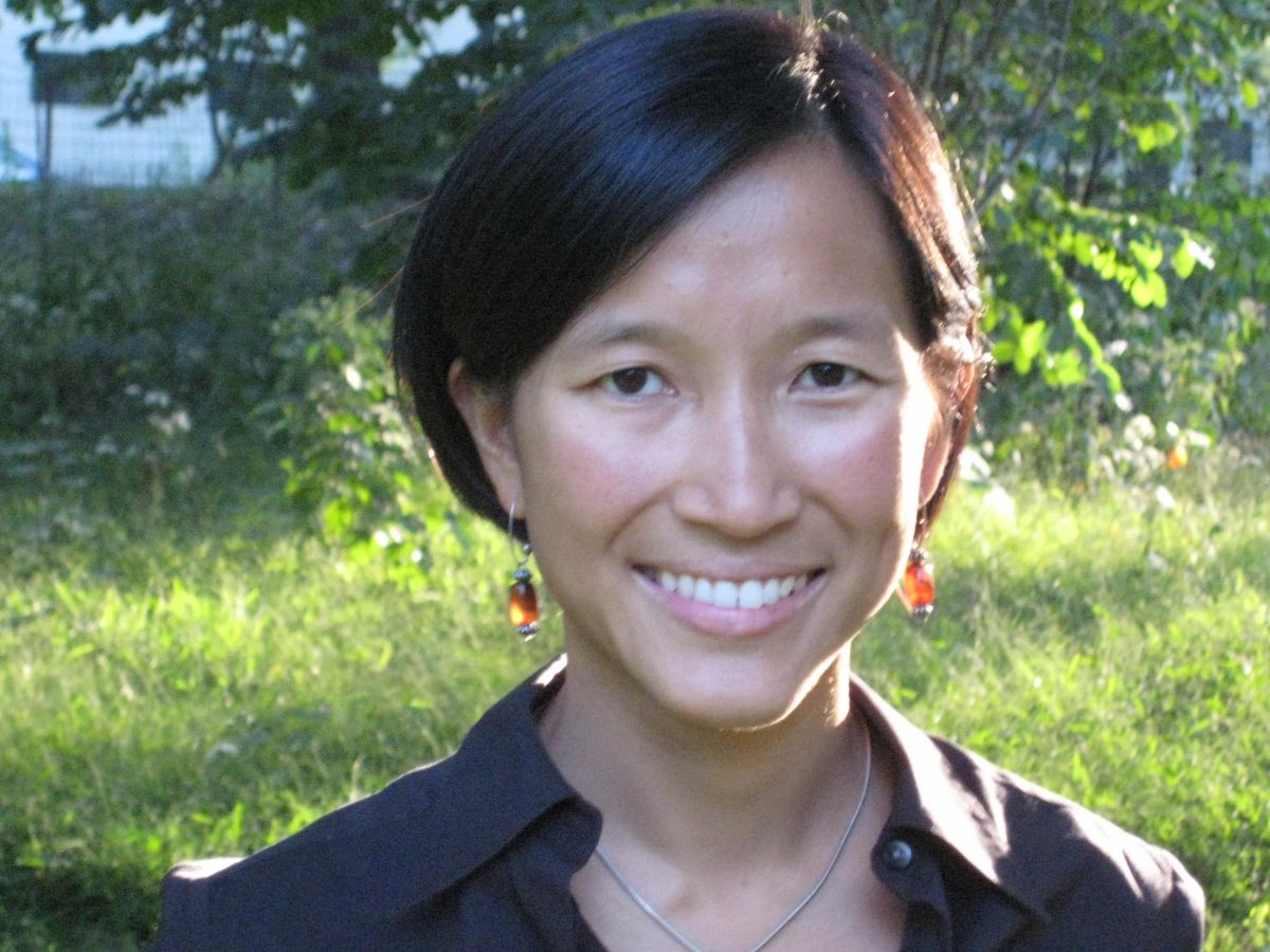 A woman with short black hair, wearing dangling earrings and a dark collared shirt, smiles outdoors with greenery and sunlight in the background.