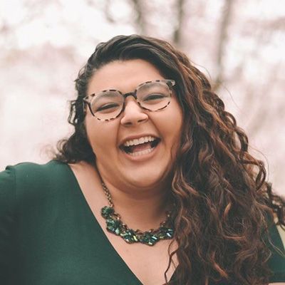 Woman with glasses and green necklace smiling warmly, wearing green top against blurred background