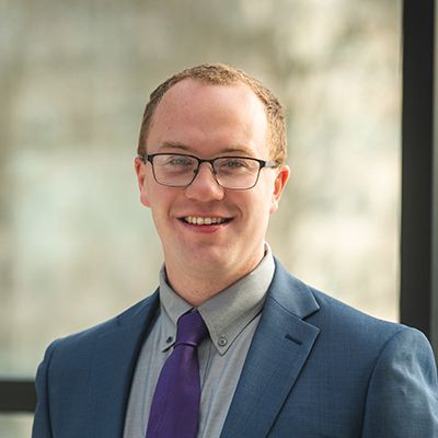 Professional headshot of smiling man in blue suit with purple tie and glasses