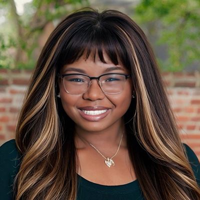 Smiling woman with long highlighted hair, glasses, and a necklace against a brick wall background