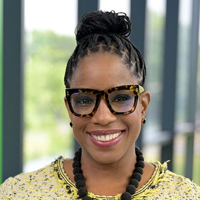 Smiling woman with braided hair wearing large black glasses and a yellow patterned top
