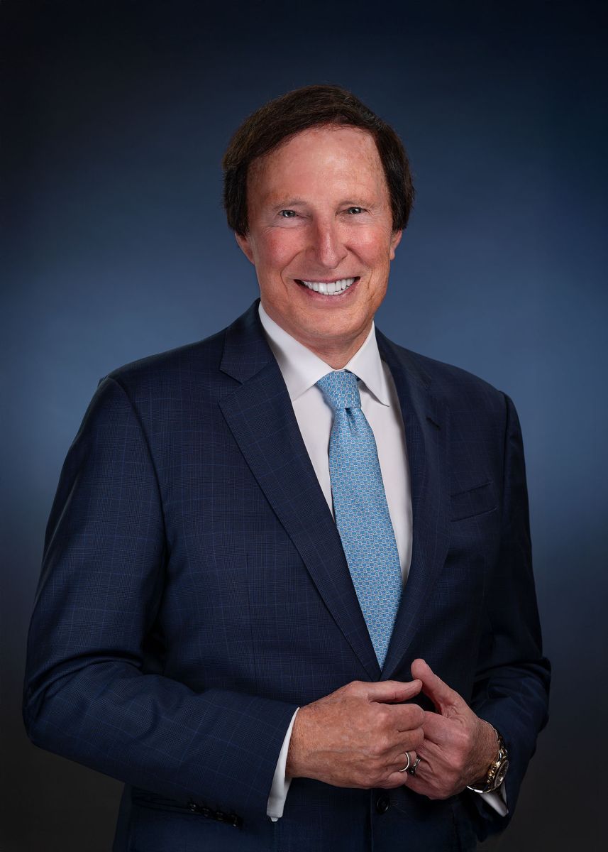 Professional headshot of smiling businessman in navy suit with blue tie against dark background