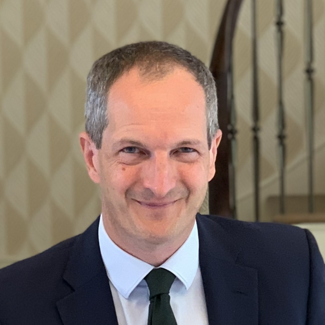 Professional headshot of a smiling man in a navy suit and tie against a beige curtain backdrop