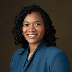 Professional headshot of a smiling woman with dark curly hair wearing a blue blazer