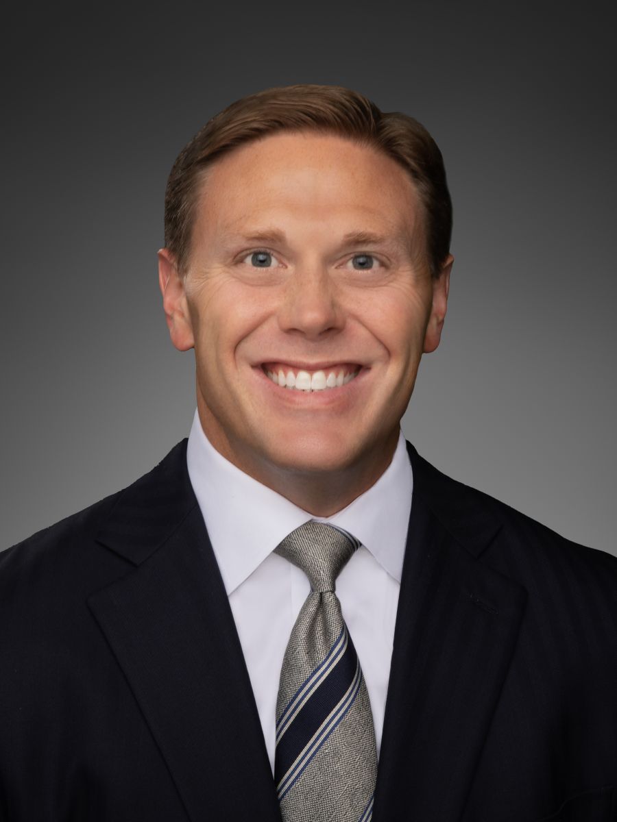 Professional headshot of a smiling man in a dark suit and striped tie against a gray background