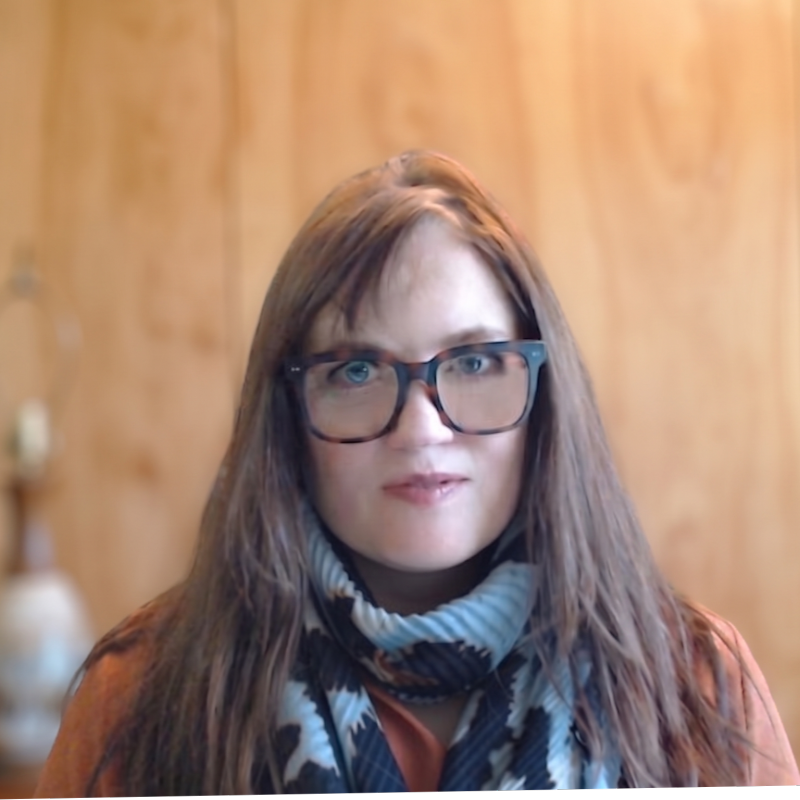 Woman with long brown hair and glasses wearing a plaid scarf against a wooden background