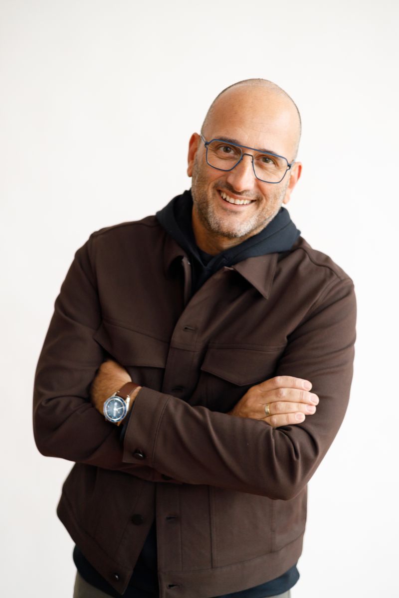 Man with glasses in dark brown jacket standing with arms crossed, smiling