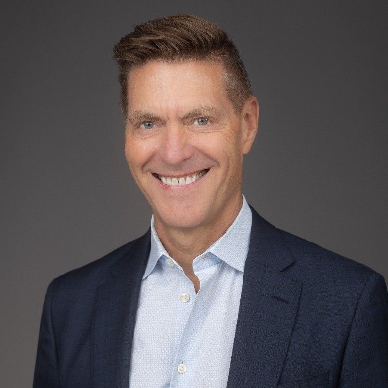 Professional headshot of smiling man in navy suit jacket and light blue shirt against dark background