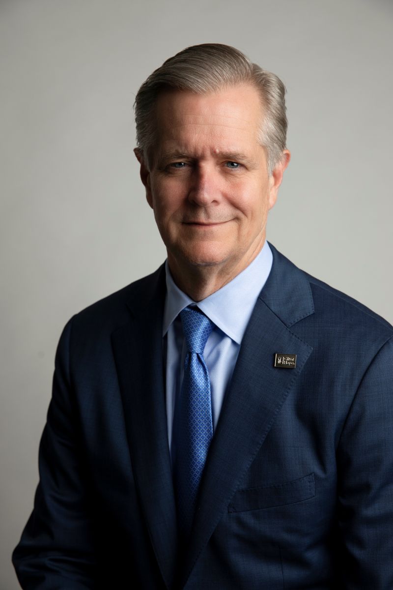 Professional headshot of a smiling man in a navy suit and blue tie against a gray background