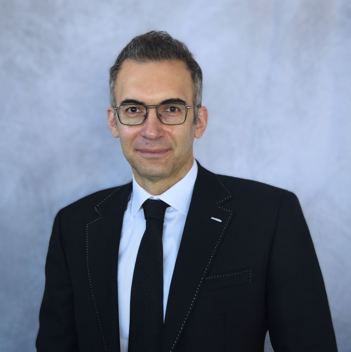Professional headshot of man in black suit and glasses with blue-gray background