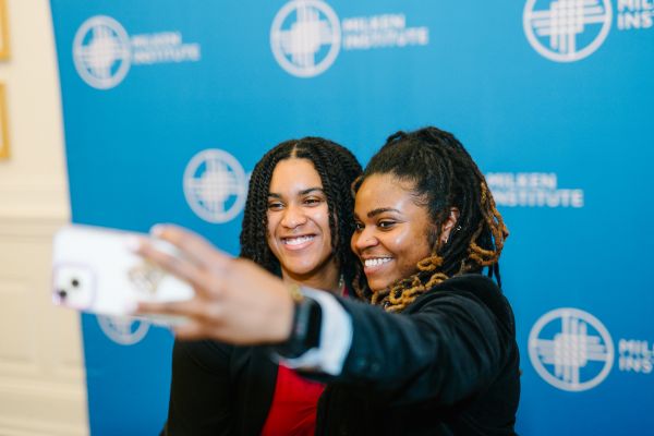 Two young Black women take a selfie in front of a step and repeat backdrop