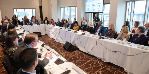 An image of several business people sitting at a panel at white tables in the shape of a rectangle, discussing the Future of Aging and Health.