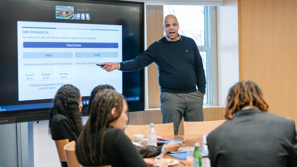 An image of HBCU 2025 Cohort Class Sessions. The Milken Institute's Inclusive Capitalism Program Insights' Team Hosts 2025 HBCU Fellows in Washington, DC. There is a diverse group of young people sitting a table taking notes while a man points at a screen and conducts a lecture.