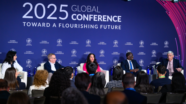 An image from the Milken Institute's Global Conference 2025 featuring five different diverse speakers sitting on a stage and having a discussion in front of an audience with a blue and pink backdrop behind them.
