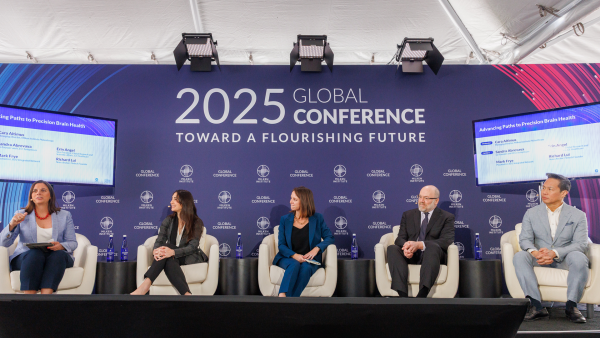 An image from the Milken Institute's Global Conference 2025. There are various diverse adult speakers sitting on a stage in white chairs and having an animated discussion. They are sitting in front of backdrop that says "2025 Global Conference, Toward a Flourishing Future."