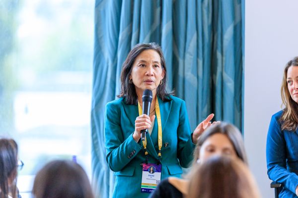 A picture of a woman who is Asian in appearance holding a microphone and speaking. She is wearing a teal blazer and has shoulder-length black hair and white skin. She is Diane Ty, managing director, Milken Institute Future of Aging (co-moderator)