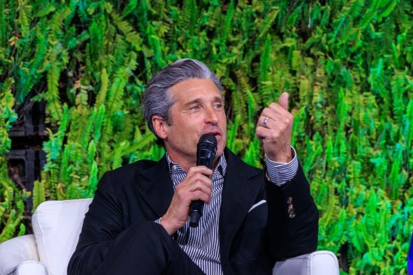 An image of Patrick Dempsey who spoke at the Milken Institute Global Conference in May 2025. He has white skin and full gray and white hair. He is speaking and sitting with greenery behind him, and wearing a dark blazer.