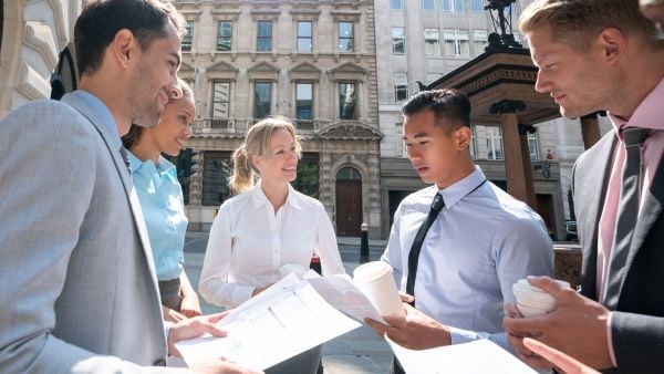 ChatGPT said: A diverse group of professionals gathered outdoors in a city setting, engaging in a collaborative discussion while reviewing documents. The group includes both men and women dressed in business attire, holding coffee cups. The background features classic architecture and city streets. The scene reflects teamwork, professional growth, and community engagement, fitting for an economic mobility alliance.