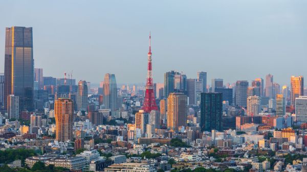 A panoramic view of Tokyo’s skyline at sunset, featuring the iconic red and white Tokyo Tower in the center, surrounded by modern high-rise buildings and skyscrapers with golden light reflecting off their glass facades.