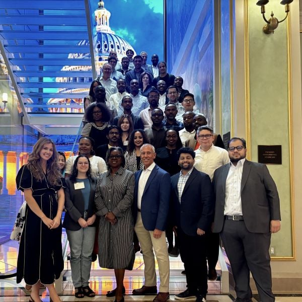 A large group of professionals pose together on a glass staircase in front of a bright digital backdrop of the U.S. Capitol dome, smiling for a formal group photo.