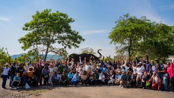 A large group of people cheerfully raise their hands outdoors in front of trees and mountains, with an elephant visible in the background.