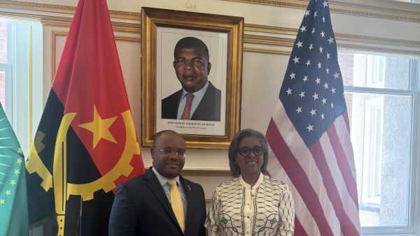 Two people stand indoors between the flags of Angola and the United States, smiling for a diplomatic-style photo beneath a framed portrait of João Manuel Gonçalves Lourenço.