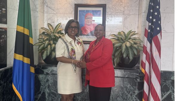 Two women stand side by side in front of the Tanzanian and U.S. flags, smiling warmly and clasping hands for a formal photo.