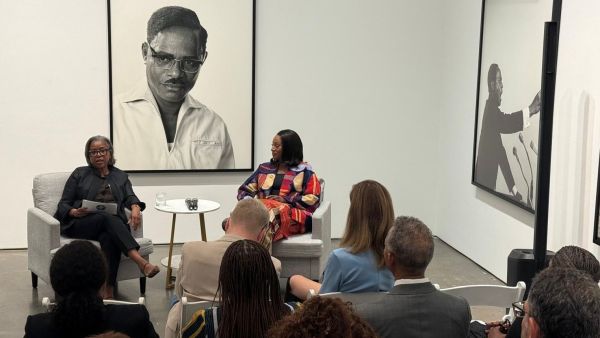 Two women sit at the front of a gallery space in conversation before an audience, framed by large black-and-white portraits of historic figures on the walls.