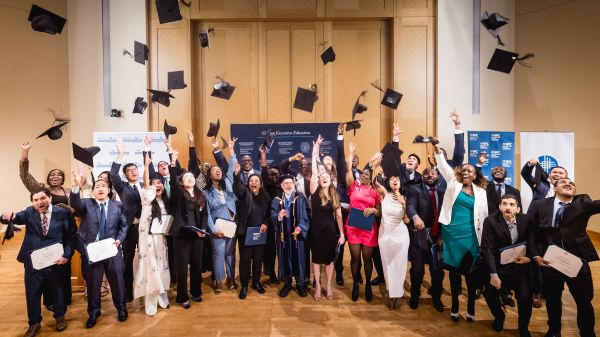 Graduates in caps and gowns celebrate together on stage, joyfully tossing their graduation caps into the air while holding diplomas and certificates.