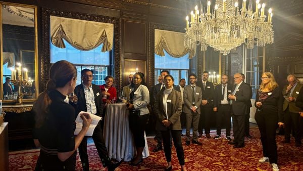 A woman speaks to a group of professionals in a richly decorated room with chandeliers, red carpet, and gold-framed mirrors, as attendees listen attentively during a networking event.
