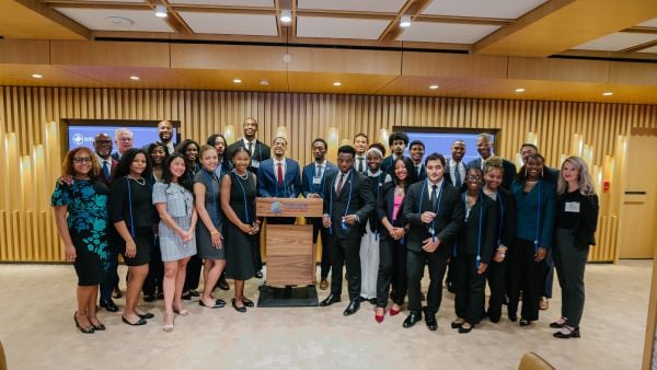 A large group of professionally dressed individuals poses for a photo in a modern conference room with wood paneling and warm lighting. They stand behind and beside a podium displaying the Milken Center for Advancing the American Dream logo. Many wear blue honor cords, and everyone smiles proudly, suggesting a celebratory or graduation event.