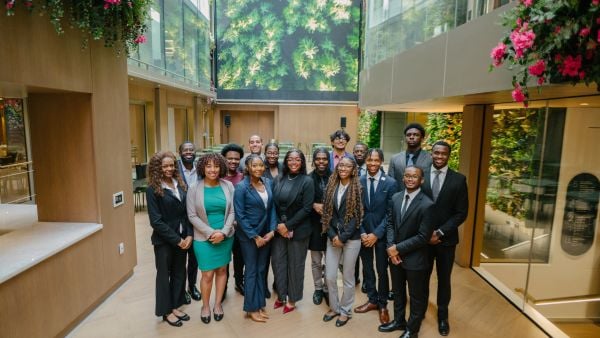 A  group of professionally dressed individuals stands together in a modern indoor atrium with wood paneling, glass walls, and lush vertical gardens. They are smiling and posing for a group photo, surrounded by greenery and natural light streaming from above.