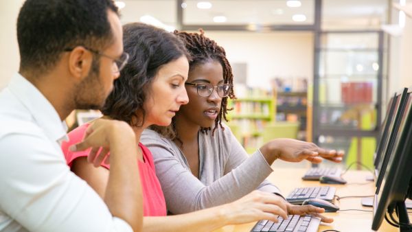Three colleagues working together at computers in an office or classroom. A woman in glasses gestures toward a monitor while explaining something to the two others, who are focused on the screen. The setting appears collaborative and instructional, with bookshelves and modern decor in the background.