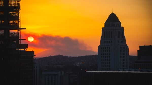 Los Angeles City Hall silhouetted at sunset as wildfire smoke drifts across the skyline, with an orange sky and the sun partially obscured.