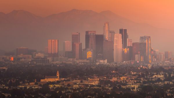 Los Angeles skyline at sunset with downtown buildings silhouetted against hazy mountains.