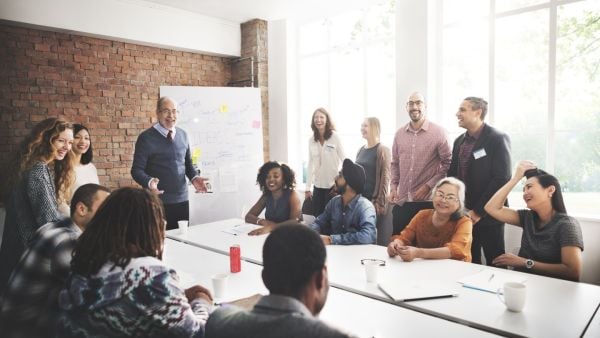 A group of adults gathered around a conference table, smiling and laughing during a collaborative meeting in a bright room with a whiteboard.