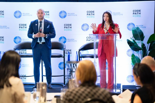 Two presenters, a man in a blue suit and a woman in a red suit, speak on a stage at a Milken Institute and CodePath.org event.