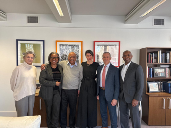 A group of six diverse professionals posing for a photo in a bright office with framed artwork and a bookshelf in the background.