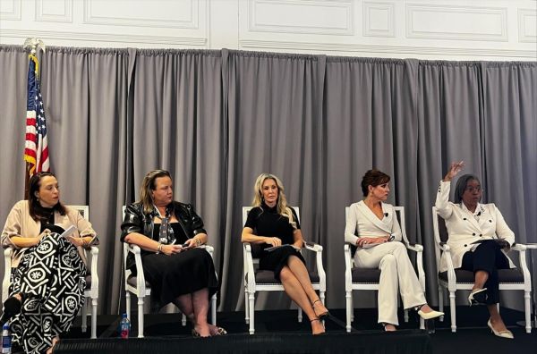 A diverse panel of five women in professional attire seated on a stage during a formal discussion.
