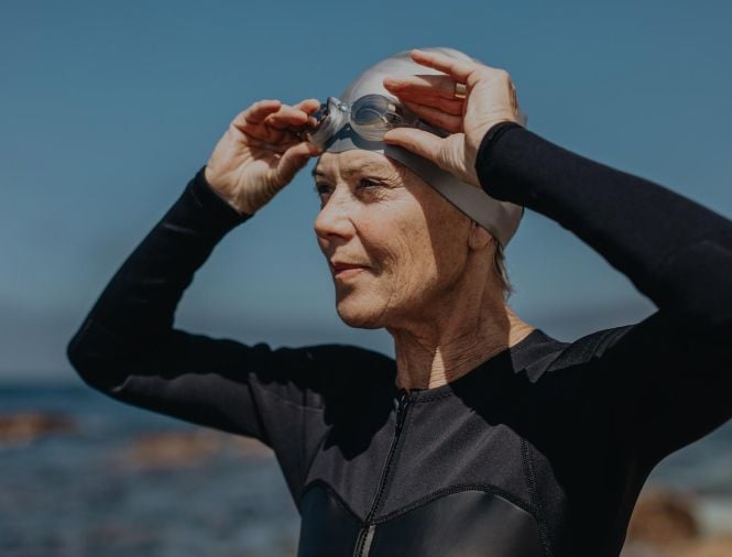 A swimmer in a wetsuit and cap adjusts their goggles while looking into the distance