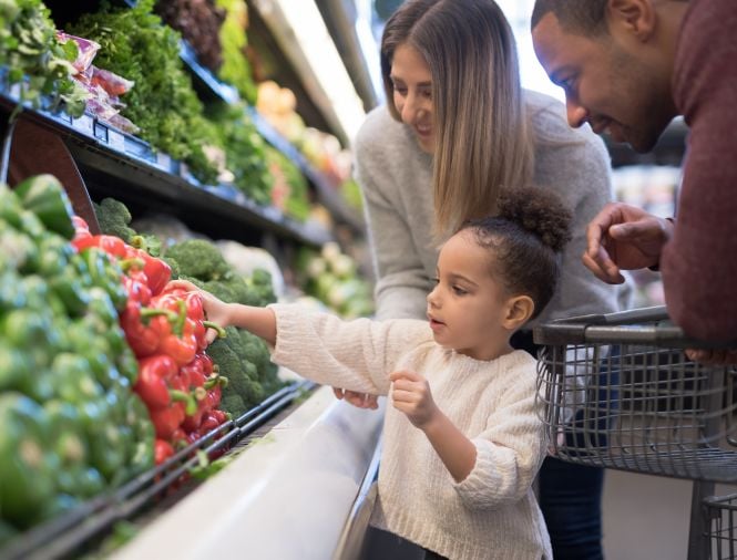 A male and female stand with young daughter in a grocery store aisle as she reaches for a red pepper in the produce section