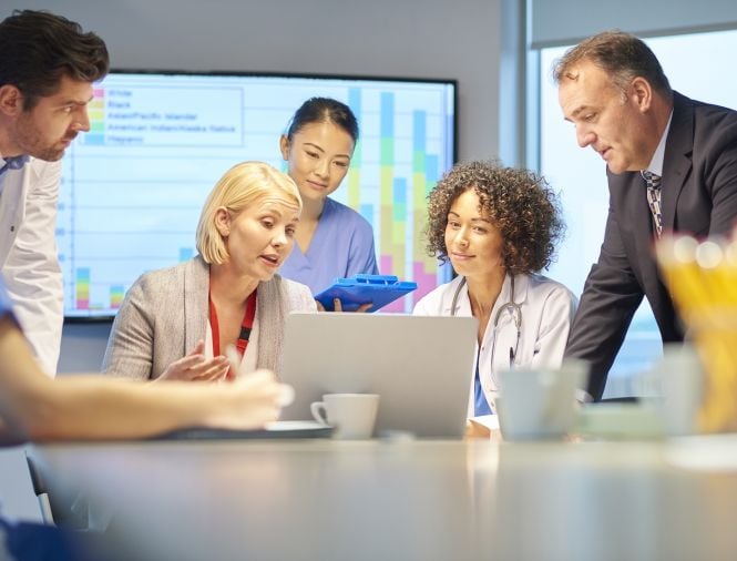 Six individuals sit around a conference table discussing something on a laptop screen in front of them