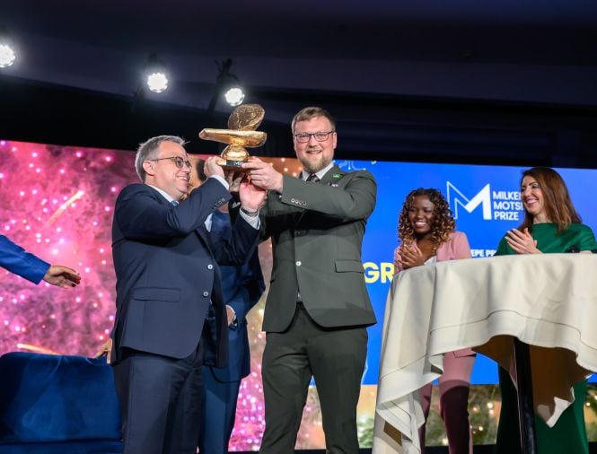 Two light-skinned males receive an award on stage at an event as two women, one Black and one white applaud for them