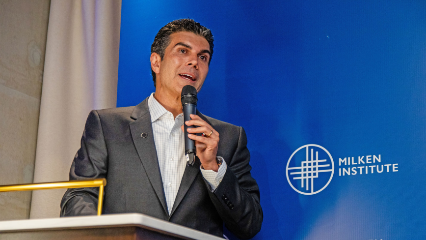 And image of a man with tan white skin and black/gray hair, holding a microphone and speaking on a stage, wearing a gray suit. He is speaking at the Milken Institute's Global Dinner Dialogues in Brazil and is standing in front of a blue "Milken Institute" background.