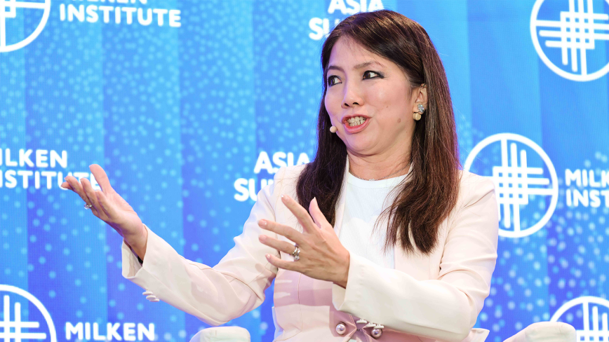 An image of Ms Tan Su Shan, the Chief Executive Officer and Director of DBS Group. She is speaking at the Milken Institute's Asia Summit 2024, and she is mid-speech with her hands gesturing. She is Asian in appearance with white skin, shoulder-length dark brown hair, and brown eyes. She is wearing a cream colored blazer over a white shirt and standing in front of a Milken Institute blue backdrop.